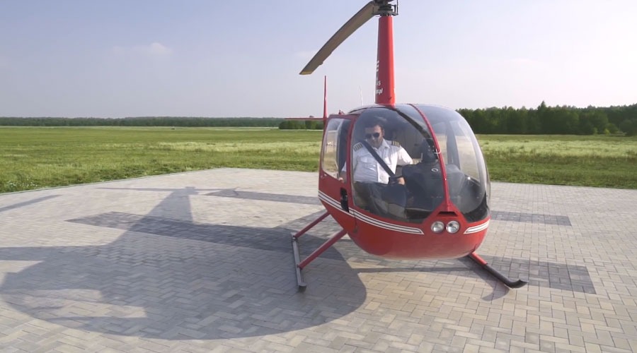 Czerwony helikopter Robinson R44 z pilotem w kokpicie, stojący na betonowym lądowisku z widocznym cieniem śmigła, w tle zielone pole i las.