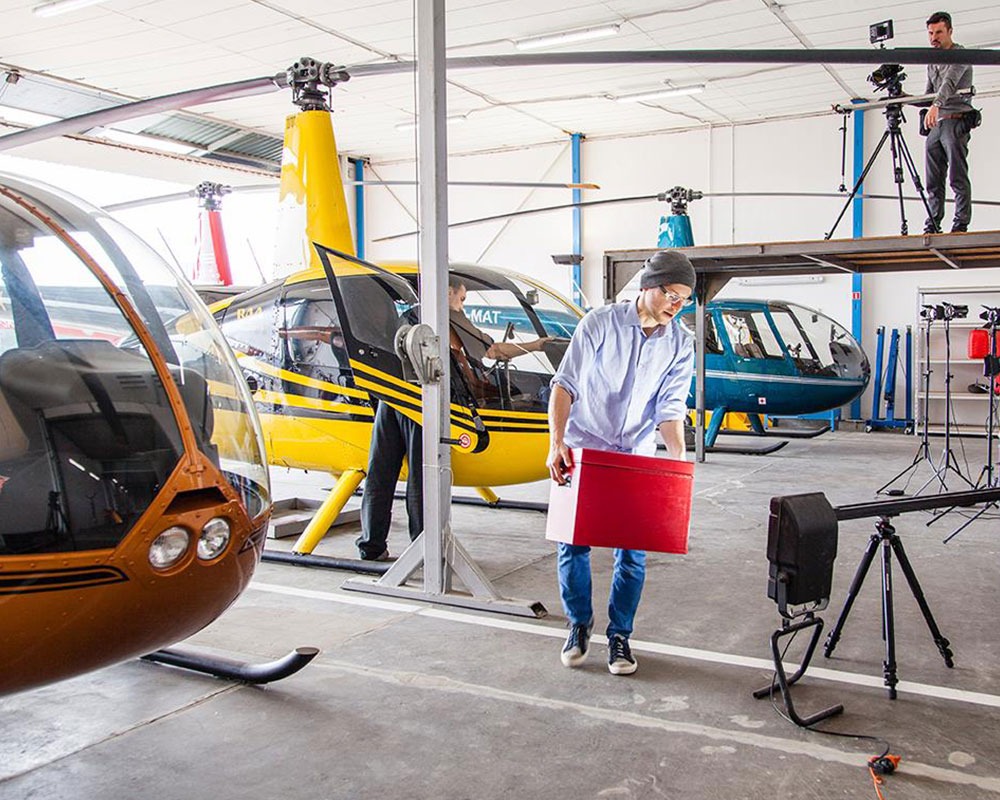 Ekipa filmowa w hangarze podczas nagrywania ujęć z helikopterami w tle, operator kamery na podwyższeniu, oświetlenie i rekwizyty rozstawione.