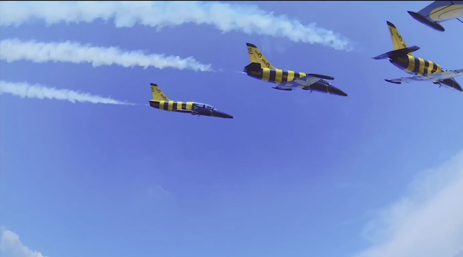 Formacja samolotów odrzutowych w locie, pomalowanych w żółto-czarne pasy, pozostawiających za sobą smugi dymu na tle błękitnego nieba.