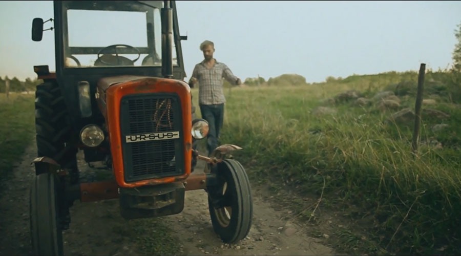 Pomarańczowy traktor Ursus na polnej drodze, w tle mężczyzna w kraciastej koszuli i zielone pole.