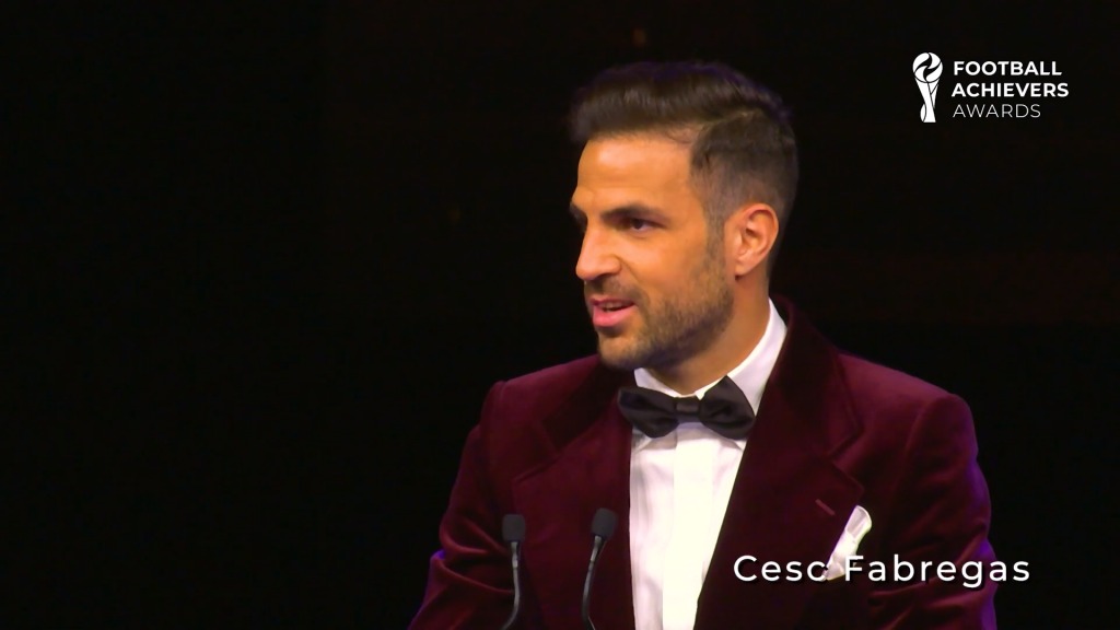 Cesc Fabregas w bordowej marynarce i muszce, przemawia podczas gali Football Achievers Awards, widoczne mikrofony i logo wydarzenia.