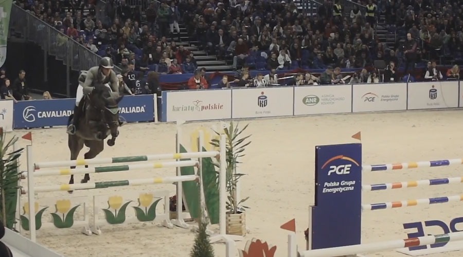 Jeździec na ciemnym koniu w trakcie skoku przez przeszkodę na zawodach Cavaliada, widownia w tle.