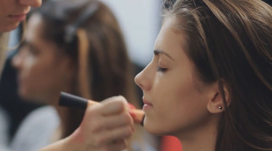 Wizażystka nakłada pędzlem podkład na twarz modelki o profilu z kolczykiem w uchu, w tle rozmyta postać.