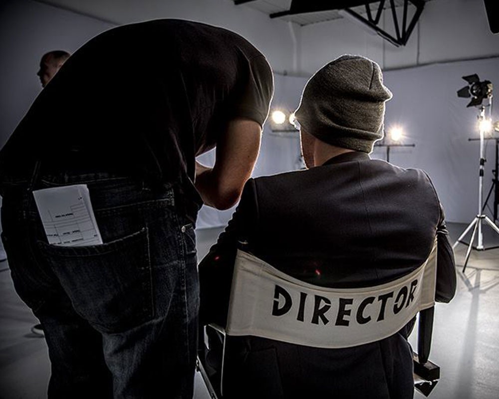 Reżyser siedzący na krześle reżyserskim na planie filmowym, oświetlenie studyjne w tle, asystent pochylony nad nim z dokumentem w kieszeni dżinsów.