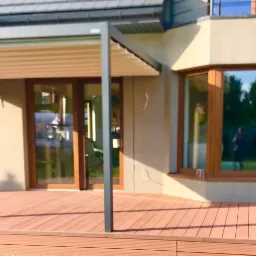 Zadaszony taras kompozytowy w kolorze brązowym przylegający do domu z dużym oknem wykuszowym i dwuskrzydłowymi drzwiami tarasowymi.