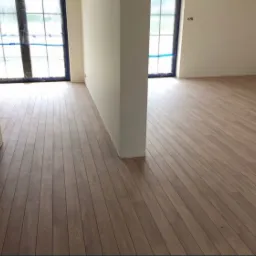 Nowo ułożona podłoga z paneli drewnianych w jasnym odcieniu brązu, widok z niskiej perspektywy, z fragmentem schodów po lewej stronie i oknami zabezpieczonymi niebieską folią w tle.