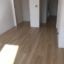 Jasne panele podłogowe w trakcie układania w surowym wnętrzu, widoczne przygotowane otwory drzwiowe i wystające kable elektryczne.