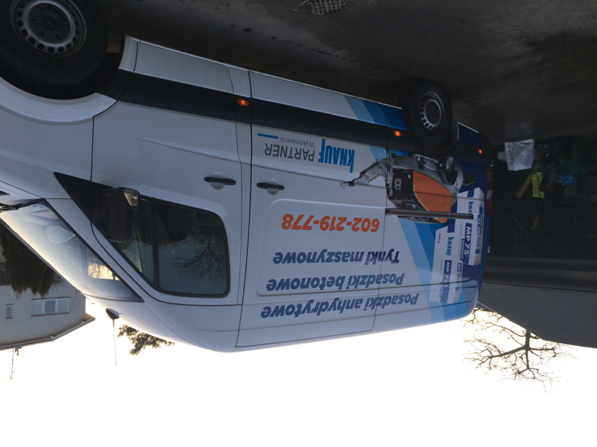 Biały samochód dostawczy z reklamą usług: posadzki anhydrytowe, posadzki betonowe i tynki maszynowe, zaparkowany na ulicy.