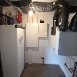 Kompletna instalacja grzewcza w kotłowni z widocznymi elementami: duży biały zasobnik, rury wentylacyjne pod sufitem, oraz skrzynki sterownicze na ścianie.