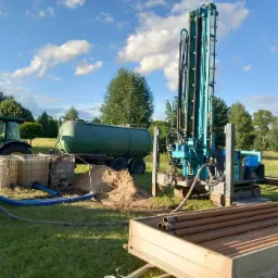 Wiertnica studni głębinowej na trawiastym terenie z traktorem i zbiornikiem na wodę w tle, widoczne rury w przyczepce i białe zbiorniki na wodę.