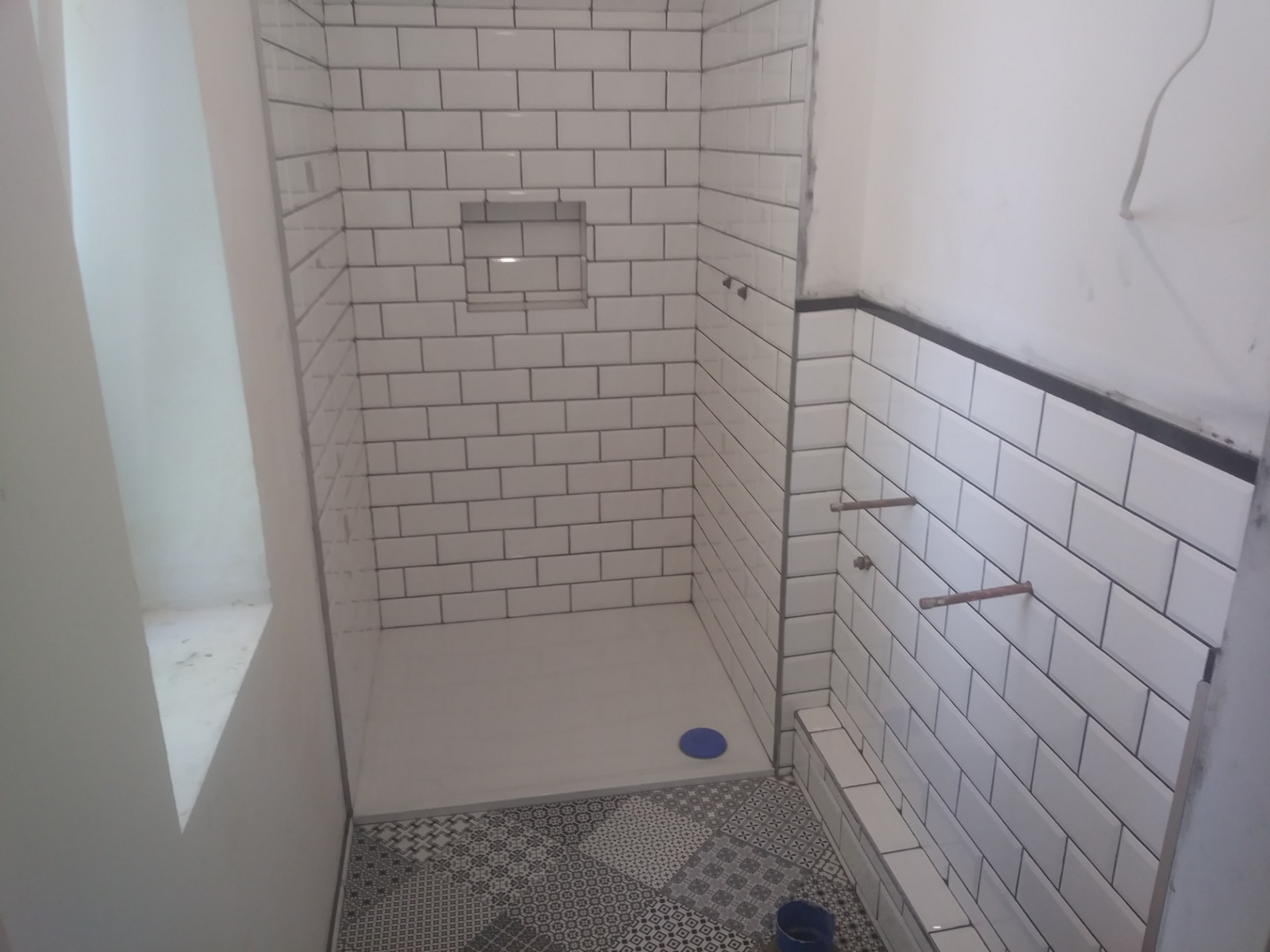 Płytki typu metro na ścianach kabiny prysznicowej z wnęką, podłoga wyłożona wzorzystą mozaiką, widoczne wystające rury instalacji wodnej.