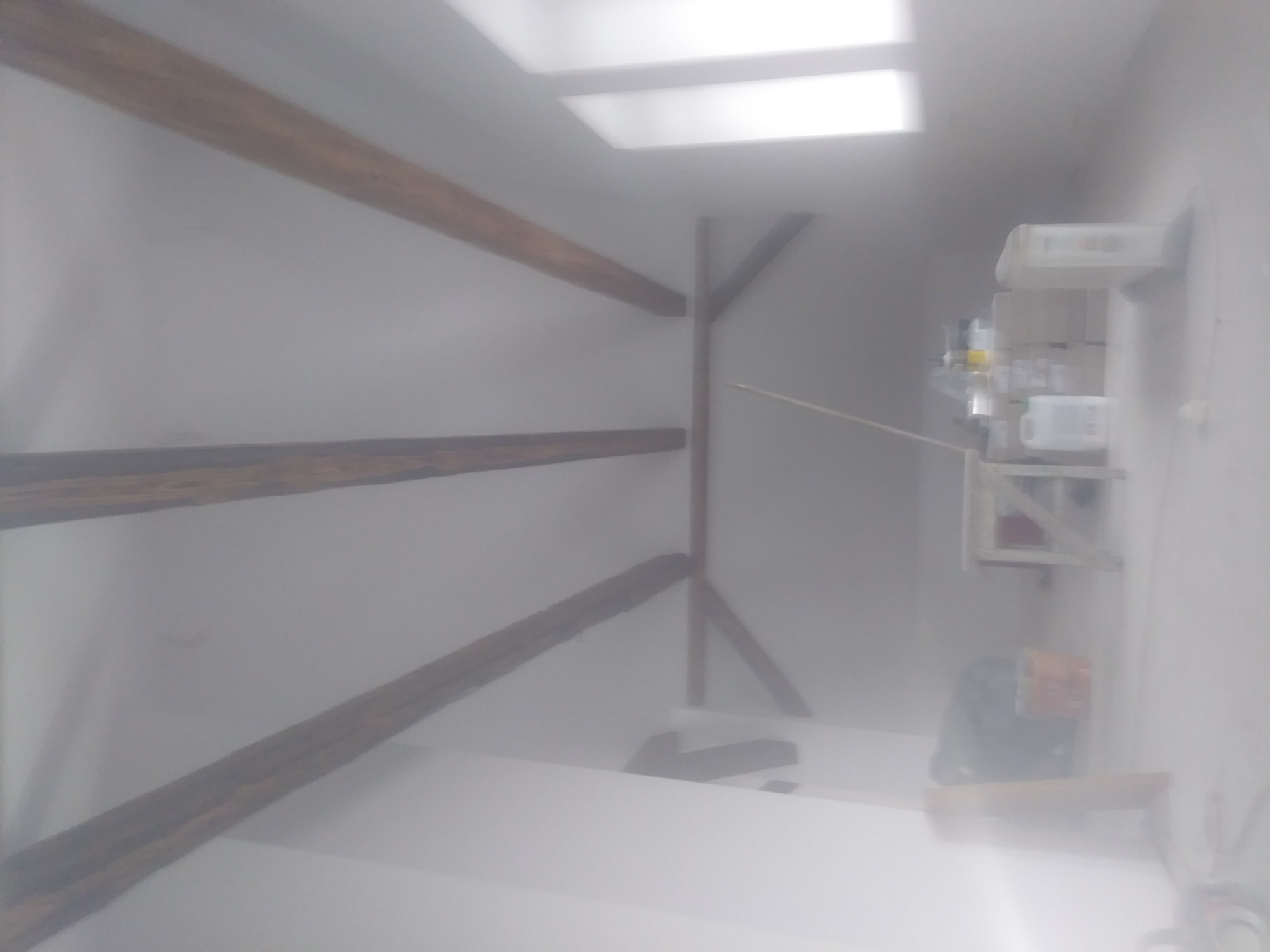 Biały sufit z drewnianymi belkami konstrukcyjnymi, widoczna półka z pojemnikami i butelkami, ujęcie z dołu w słabym oświetleniu.
