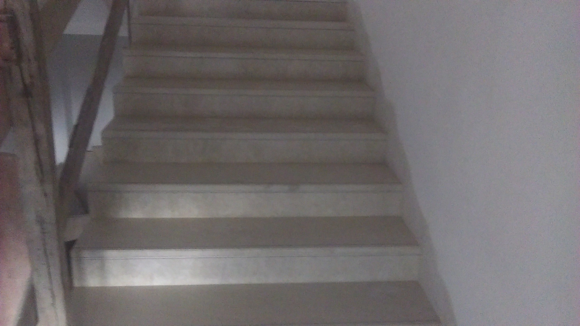 Widok z dołu na wyłożone płytkami schody z drewnianą balustradą, oświetlone z góry, z widoczną fakturą płytek.