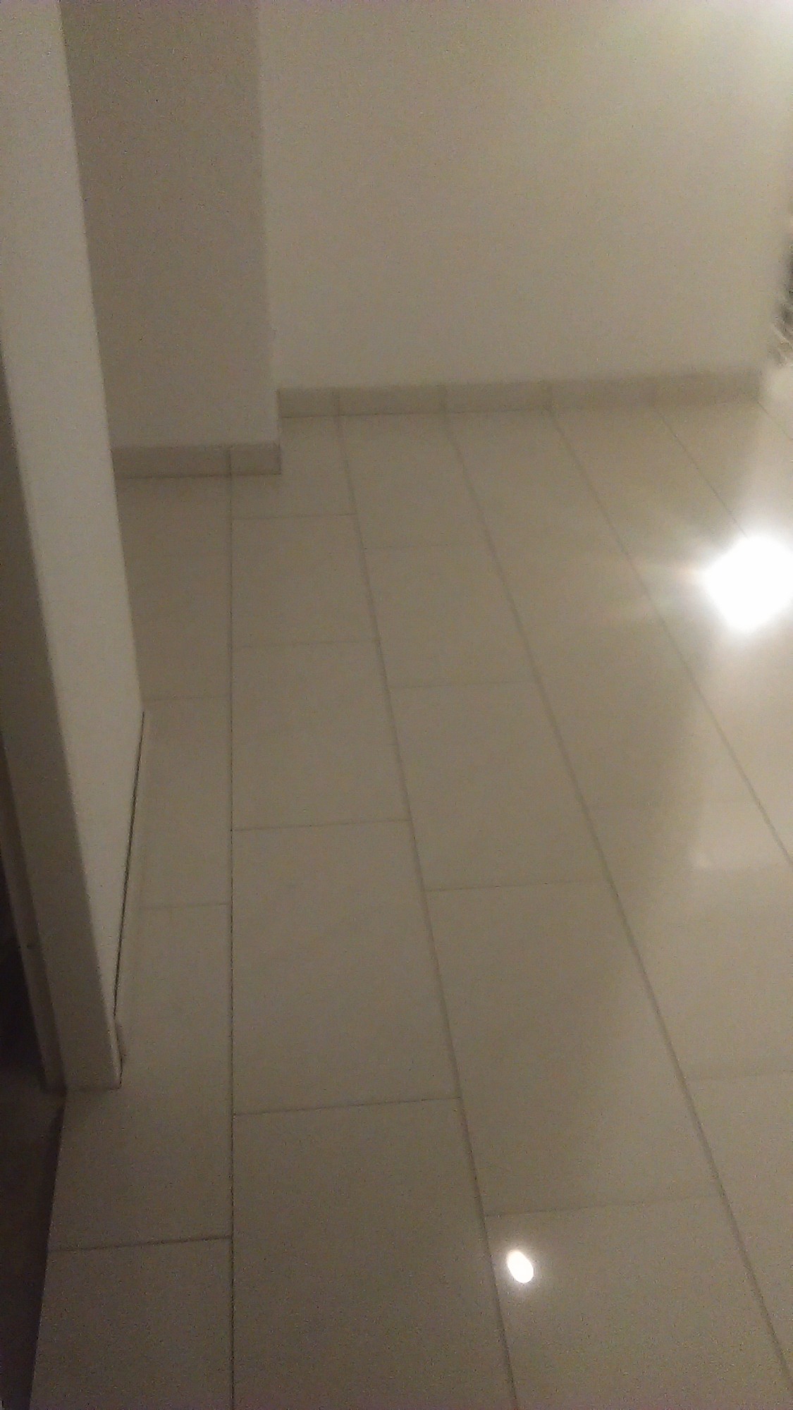 Ułożona jasna, kwadratowa glazura podłogowa z widocznymi fugami i listwą przypodłogową przy białej ścianie.