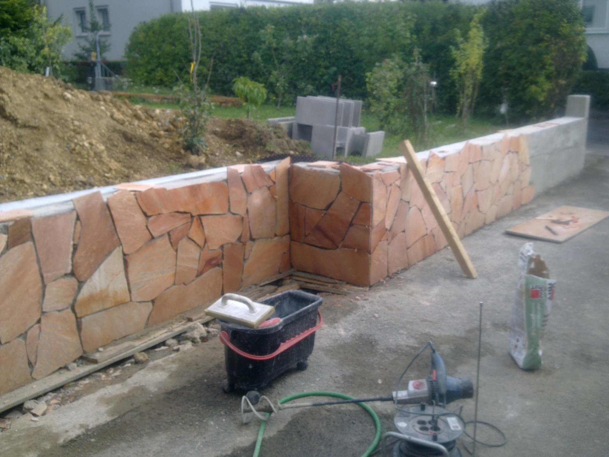 Murowana ściana z nieregularnych, brązowo-pomarańczowych kamieni, częściowo wykończona, z widocznymi narzędziami glazurniczymi i zaprawą w wiadrze na kółkach, na tle zieleni i budynków.