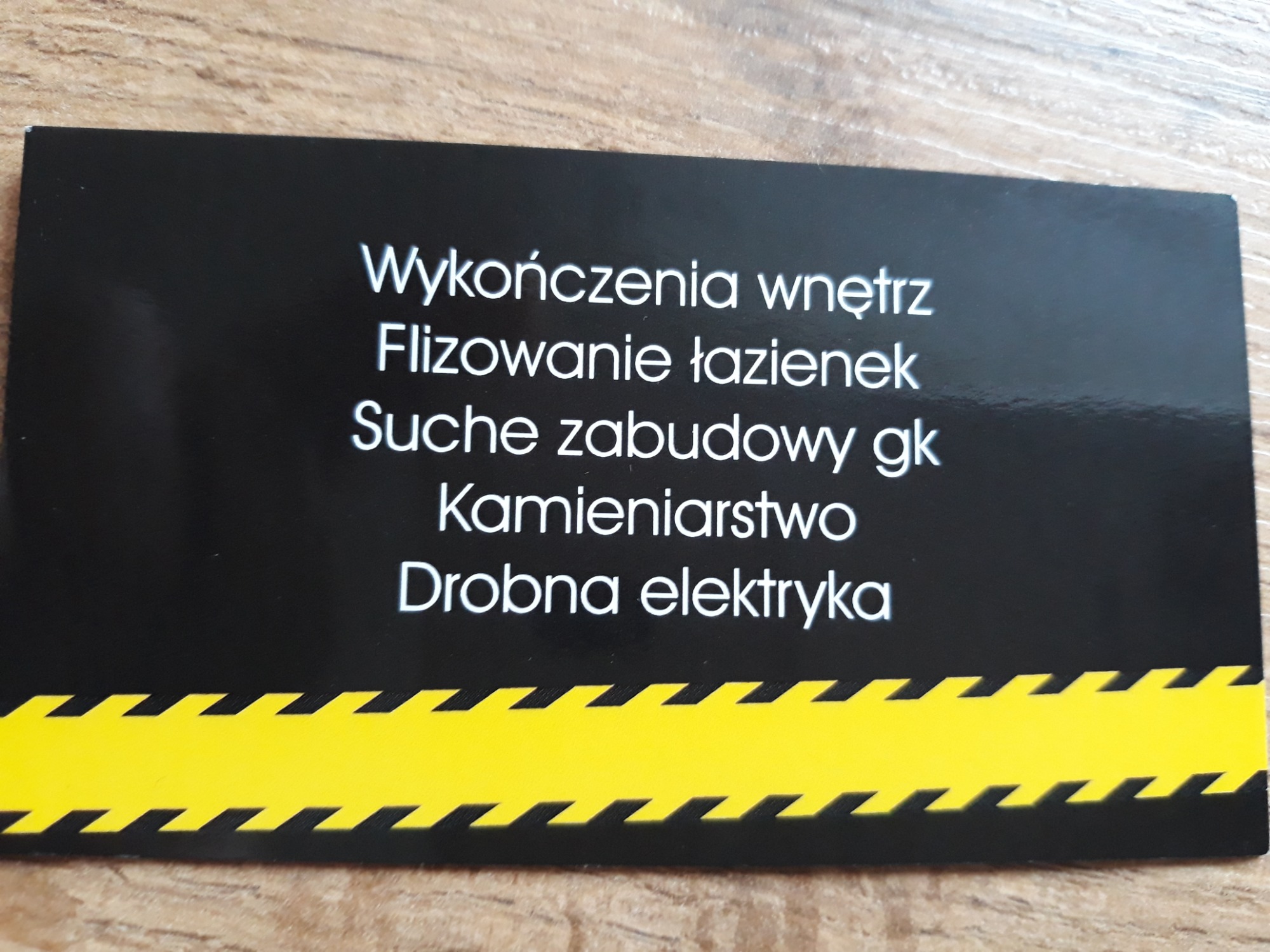 Czarna wizytówka z białym tekstem informującym o usługach wykończenia wnętrz, flizowania łazienek, suchych zabudów GK, kamieniarstwa i drobnej elektryki, z żółtym pasem z czarnymi zygzakami na dole.