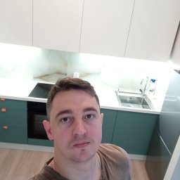 RJA Building - Selfie mężczyzny na tle nowej kuchni z białymi szafkami górnymi i zielonymi dolnymi, z drewnianą podłogą i blatem z białego kamienia.