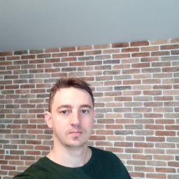 RJA Building - Mężczyzna na tle ściany z cegły dekoracyjnej. Widoczne różne odcienie cegieł i fugi. T-shirt w kolorze ciemnej zieleni.