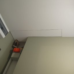 RJA Building - Fragment wnętrza z gładką, beżową ścianą i białym sufitem, widoczne detale wykończenia. Narożnik pomieszczenia z elementami wyposażenia w tle.