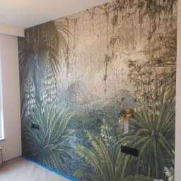 RJA Building - Ściana z fototapetą w motywy roślinne, zabezpieczona taśmą malarską przy podłodze. Widoczna lampa ścienna i gniazdka elektryczne. Dekoracja wnętrza w stylu tropikalnym.