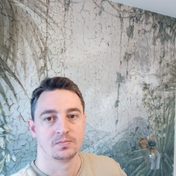 RJA Building - Mężczyzna na tle ściany z dekoracyjną tapetą imitującą spękaną farbę i motywy roślinne. Widoczny włącznik światła i ozdobny kinkiet.