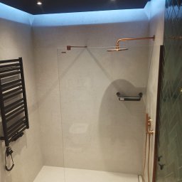 RJA Building - Nowoczesna kabina prysznicowa walk-in z miedzianą armaturą, grzejnikiem drabinkowym i oświetleniem LED w wykończonej łazience. Widoczne płytki na ścianach i podłodze.