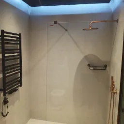 Nowoczesna kabina prysznicowa walk-in z deszczownicą w odcieniach miedzi, grzejnikiem drabinkowym i oświetleniem LED. Minimalistyczny design z jasnymi płytkami.
