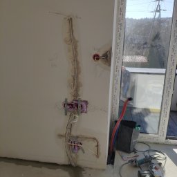 RJA Building - Ściana w trakcie prac instalacyjnych: widoczne kable, puszki elektryczne zabezpieczone pianką montażową, otwory w ścianie, narzędzia leżące na podłodze, okno balkonowe w tle.