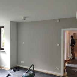 RJA Building - Pokój w trakcie malowania: szare ściany, podłoga zabezpieczona folią, odkurzacz budowlany, widoczny fragment okna i drzwi z sylwetką osoby w oddali.