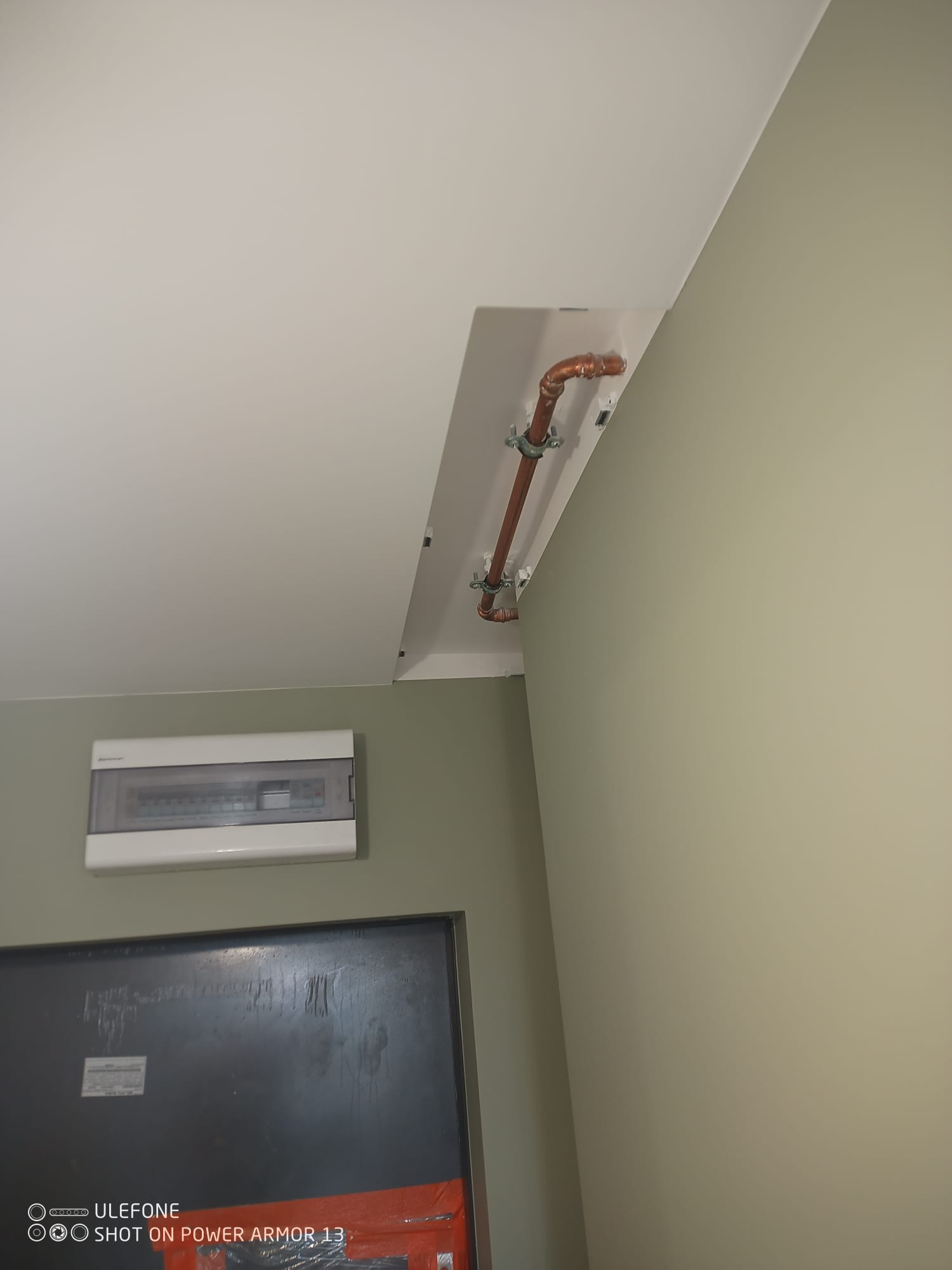 Miedziana rura pod sufitem, przymocowana do białej listwy maskującej, obok skrzynka elektryczna na zielonej ścianie. Widok z dołu, blisko drzwi.