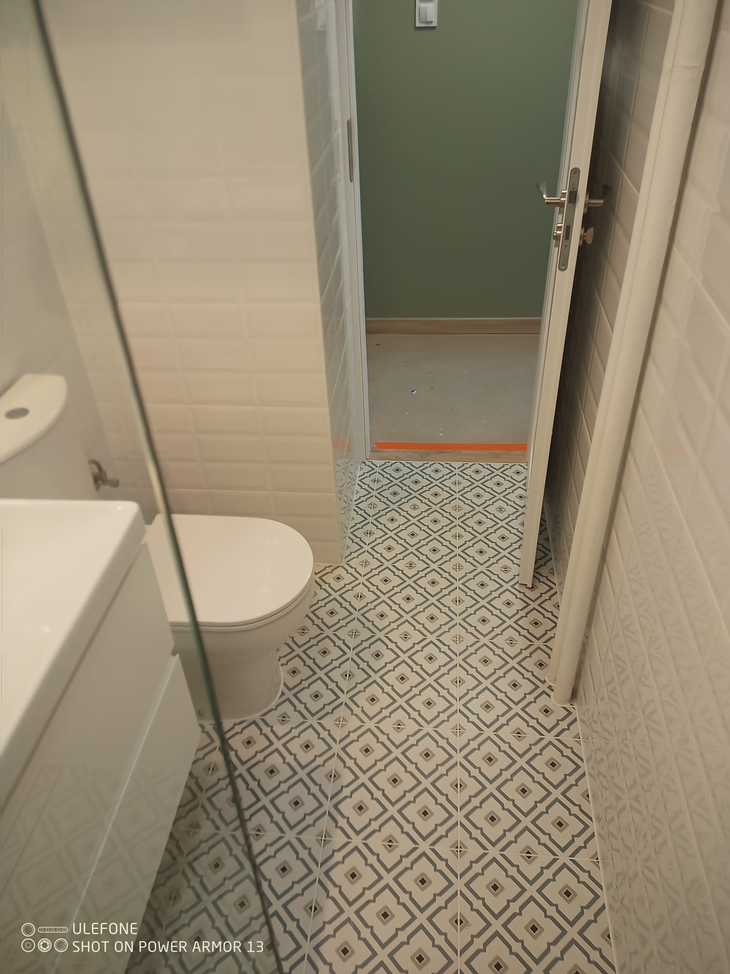 Wykończona łazienka z toaletą, umywalką i wzorzystą podłogą. Białe płytki na ścianach, widok na kolejne pomieszczenie z zieloną ścianą. Nowoczesny design.