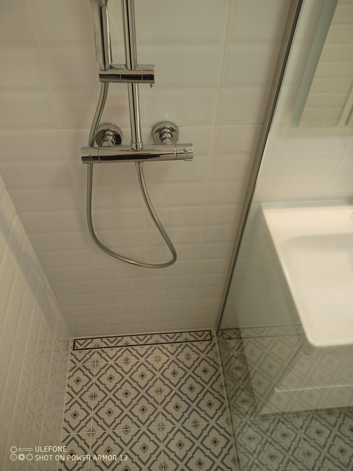 Nowoczesna łazienka z chromowaną armaturą prysznicową, jasnymi, kwadratowymi płytkami na ścianach i dekoracyjnymi płytkami podłogowymi w geometryczny wzór.