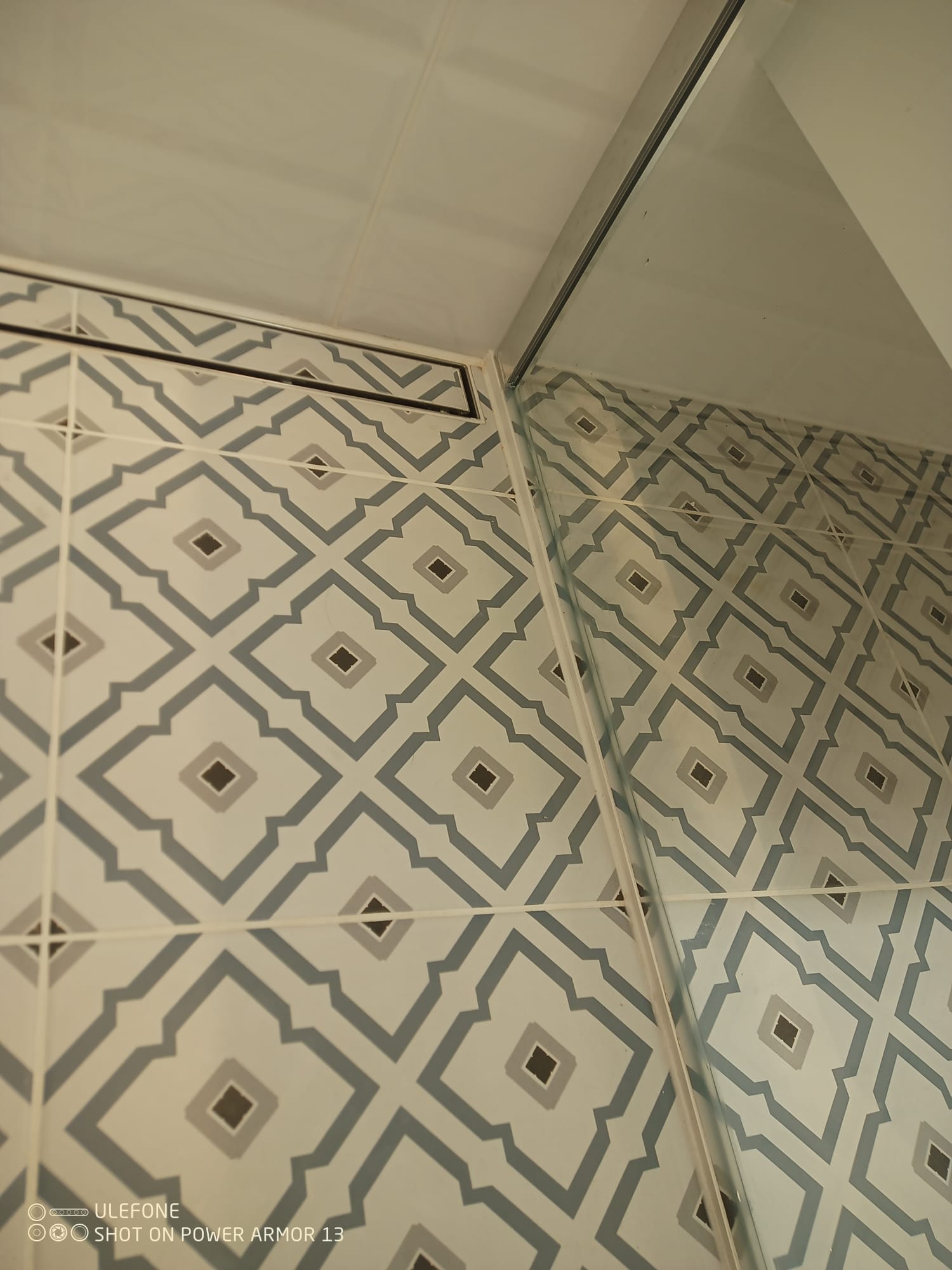 Wzorzyste płytki w łazience z odpływem liniowym i szklaną ścianką kabiny, ułożone pod kątem. Geometryczny wzór w odcieniach szarości i bieli.