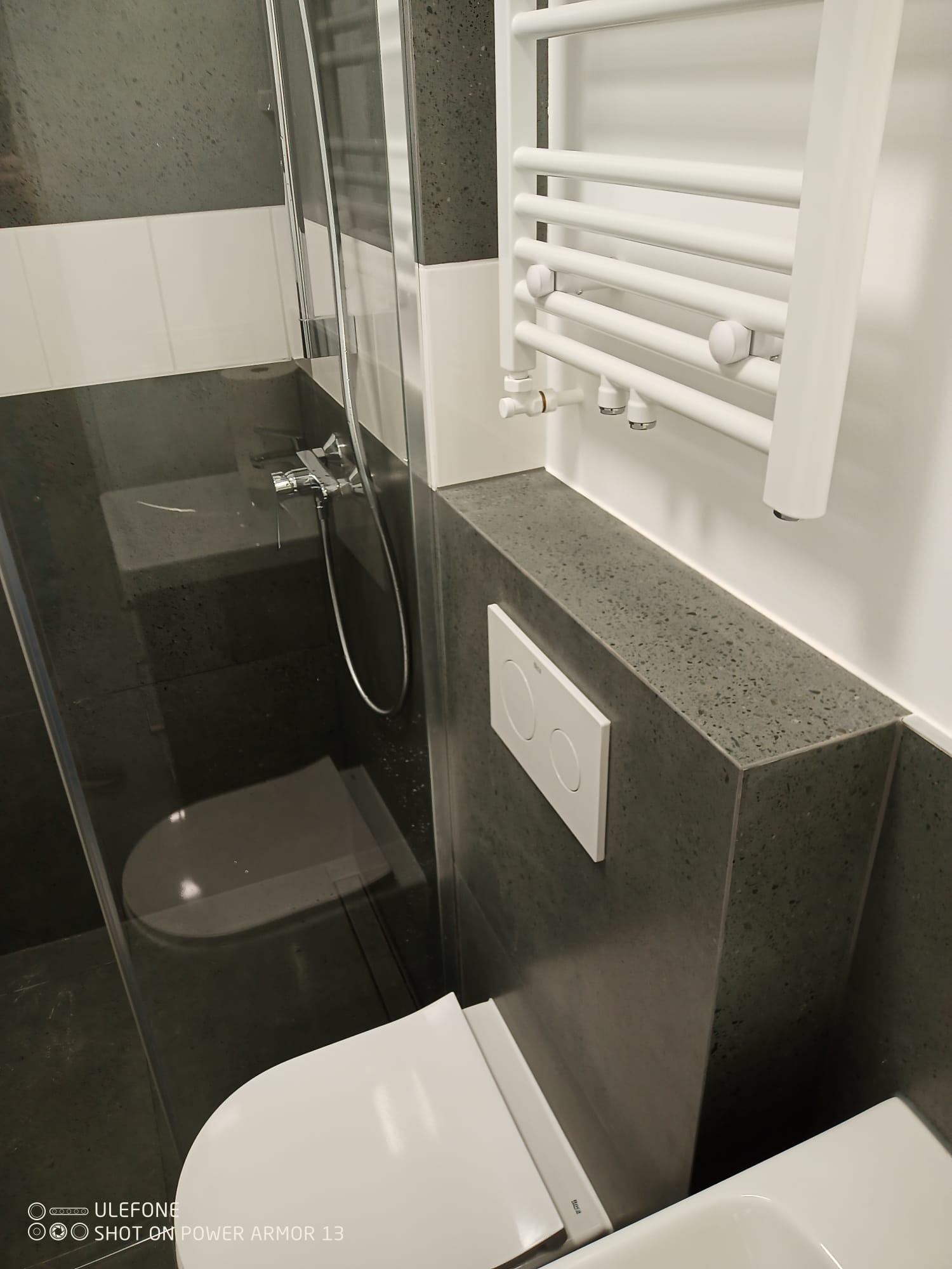 Nowoczesna łazienka w szaro-białej tonacji: kabina prysznicowa ze szklaną ścianką, podwieszana toaleta z przyciskiem i biały grzejnik drabinkowy.