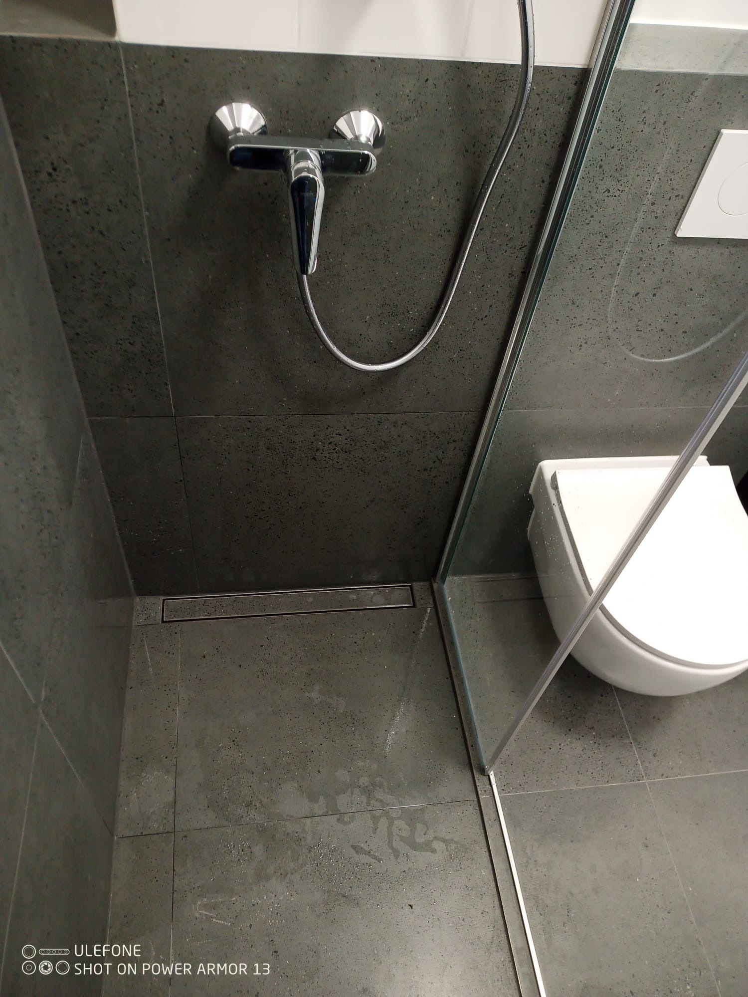 Nowoczesna kabina prysznicowa z szarymi płytkami, odpływem liniowym i szklaną ścianką. Minimalistyczny design, widoczny fragment toalety.