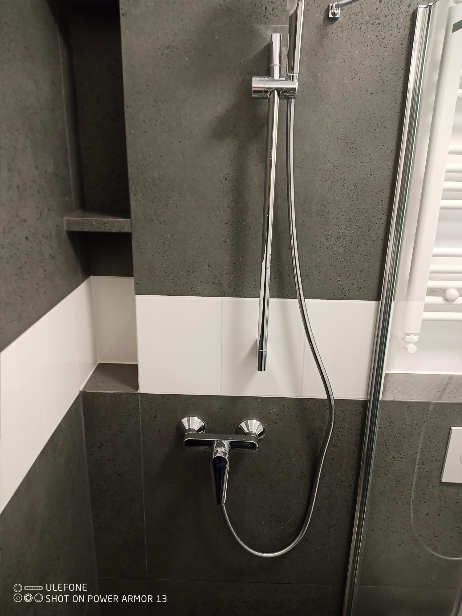 Szara ściana w łazience z chromowaną baterią i kabiną prysznicową. Widoczna półka wnękowa i biały grzejnik drabinkowy. Minimalistyczny design.