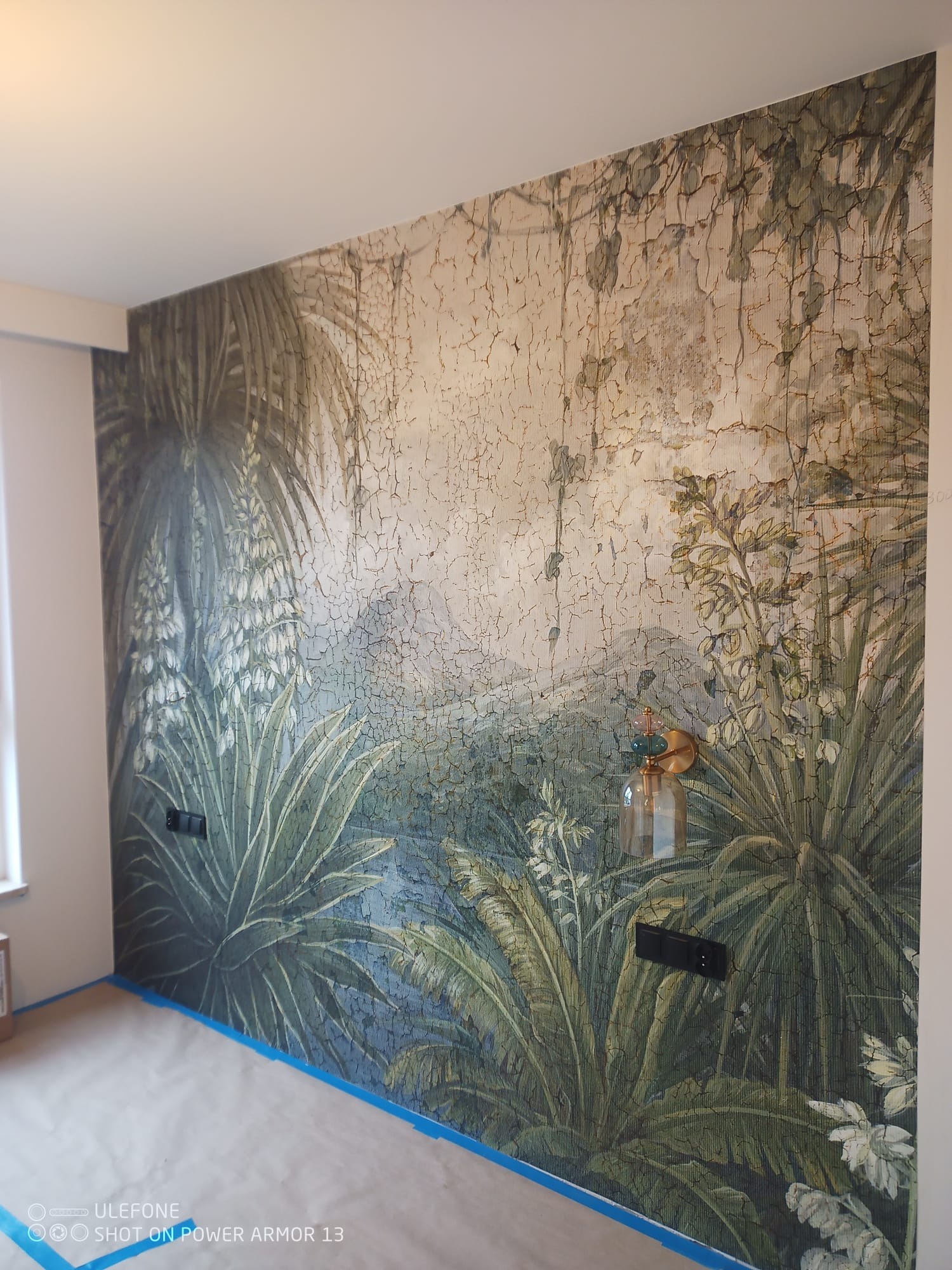 Ściana z fototapetą w motywy roślinne, zabezpieczona taśmą malarską przy podłodze. Widoczna lampa ścienna i gniazdka elektryczne. Dekoracja wnętrza w stylu tropikalnym.