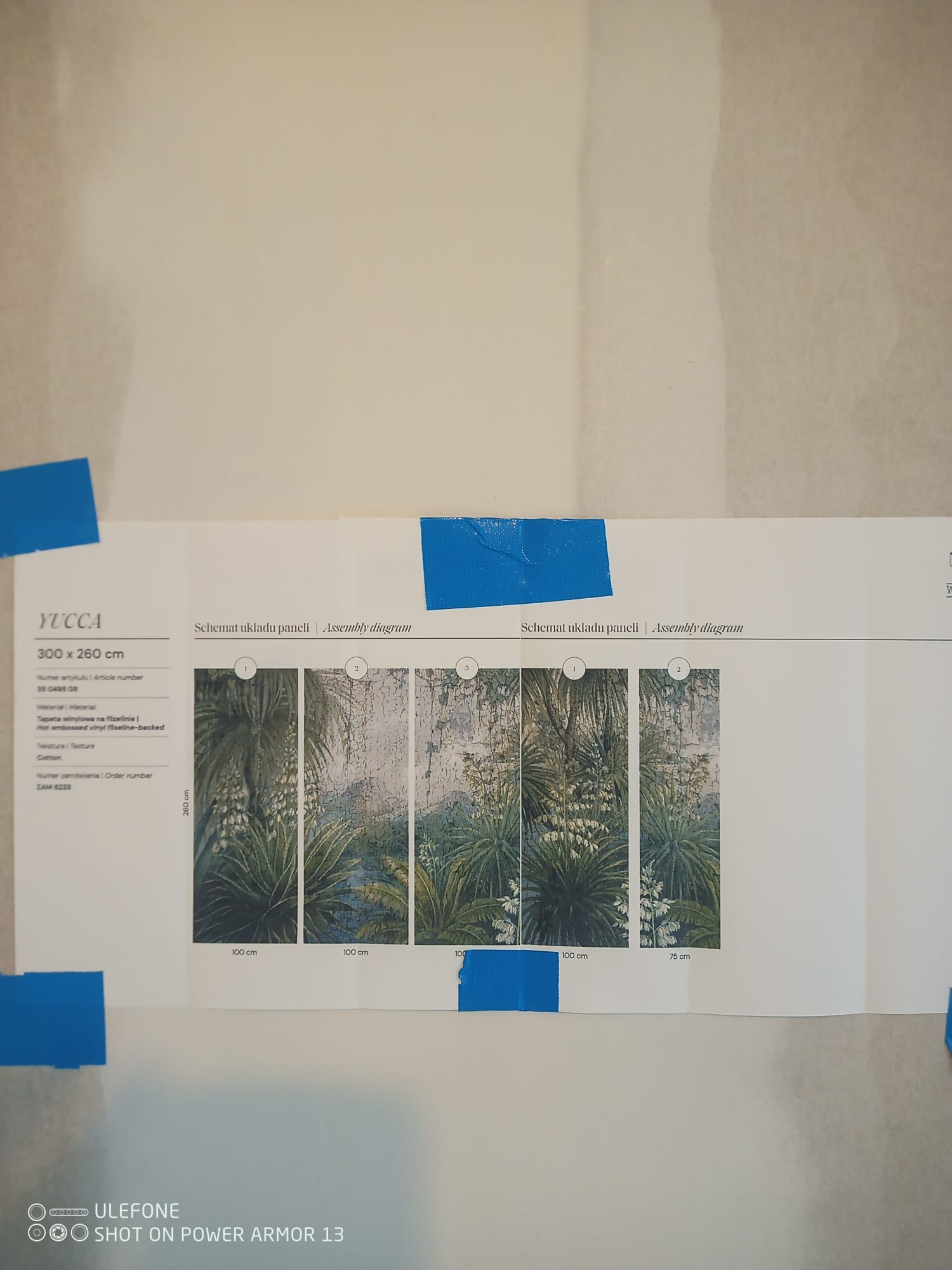 Schemat układu paneli tapety winylowej Yucca (300x260cm) z motywem roślinnym, przyklejony na ścianie za pomocą niebieskiej taśmy, prezentujący instrukcję montażu.