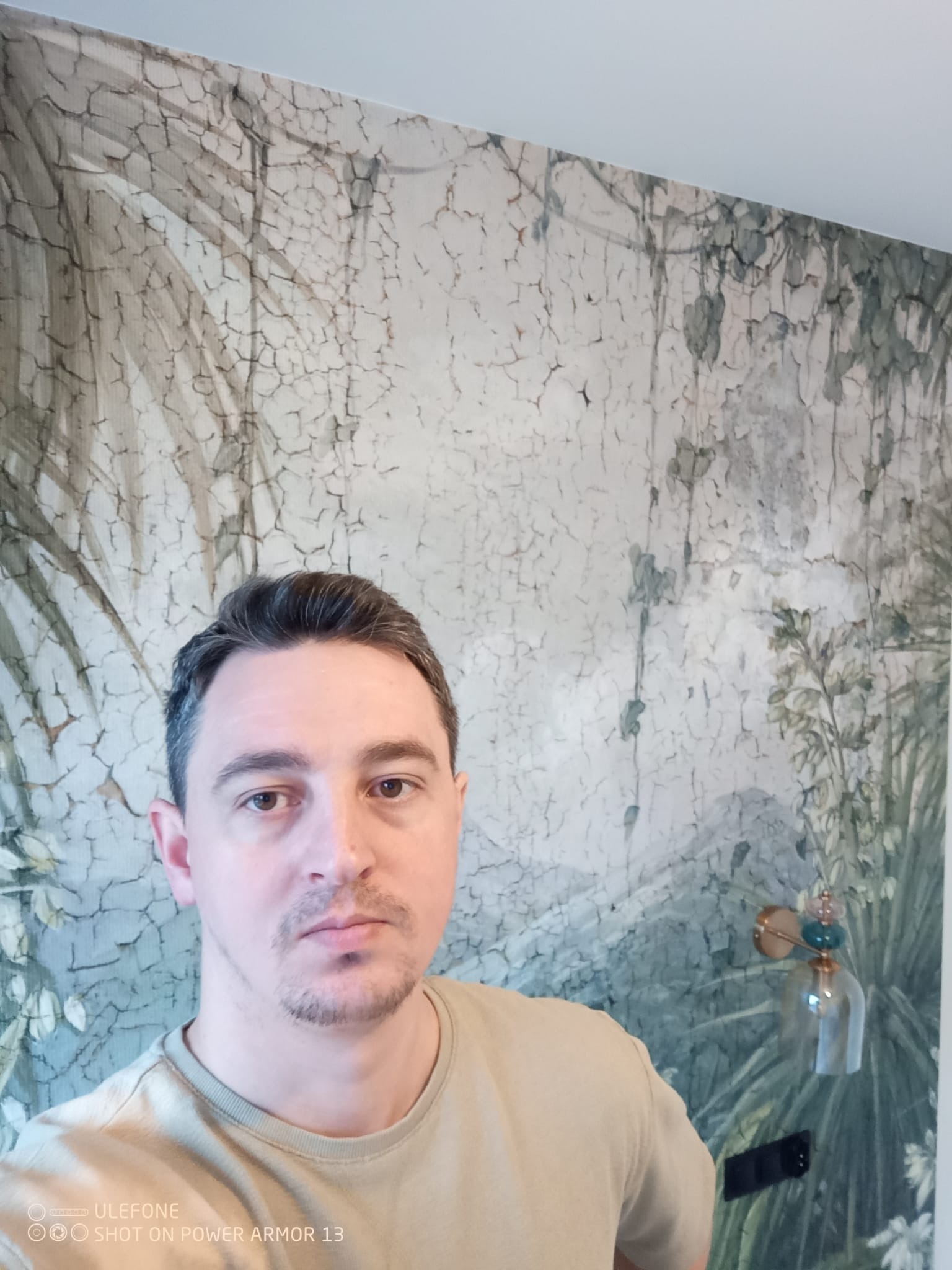 Mężczyzna na tle ściany z dekoracyjną tapetą imitującą spękaną farbę i motywy roślinne. Widoczny włącznik światła i ozdobny kinkiet.