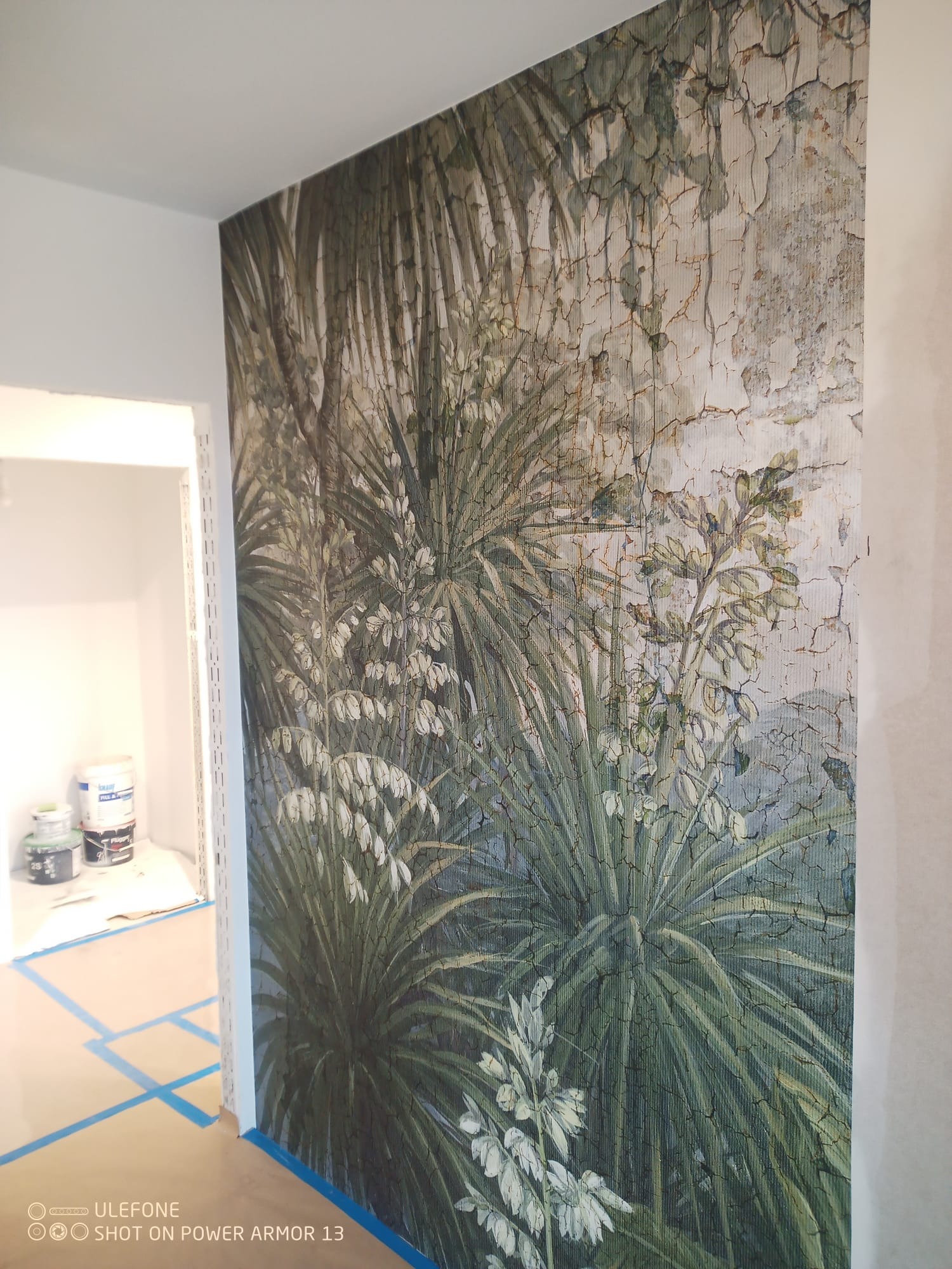 Ściana z fototapetą w tropikalne rośliny, widok z boku, podłoga zabezpieczona folią i taśmą malarską, w tle widoczne puszki z farbami.