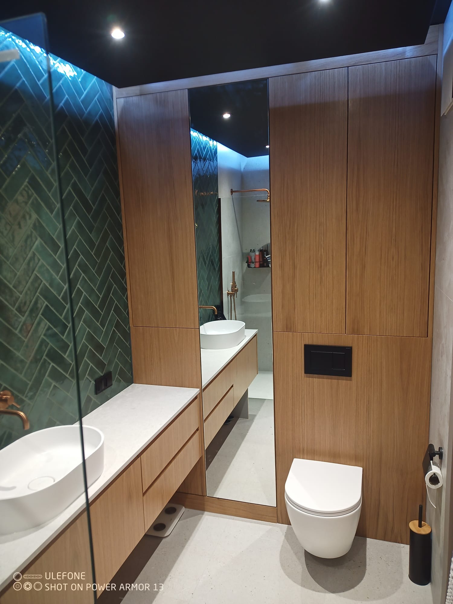 Nowoczesna łazienka z zieloną, ułożoną w jodełkę glazurą, drewnianą zabudową i białymi umywalkami. Lustro optycznie powiększa przestrzeń, a czarny sufit dodaje elegancji.