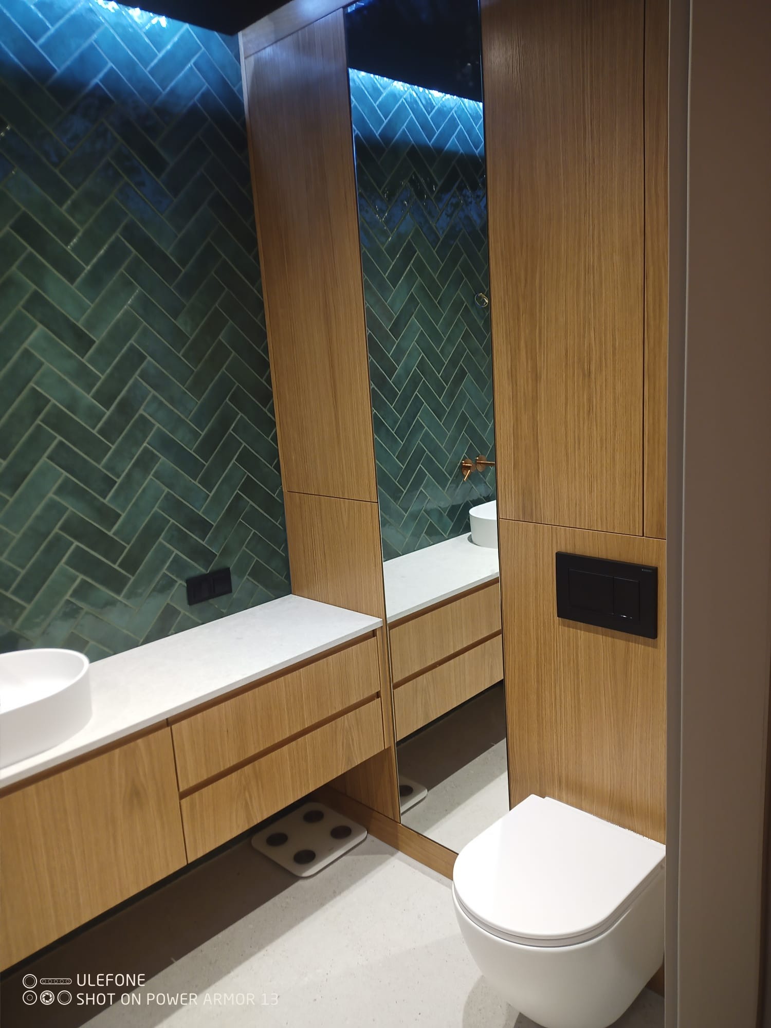 Nowoczesna łazienka z zieloną, ułożoną w jodełkę glazurą, drewnianymi szafkami pod umywalką, lustrem i podświetleniem LED. Minimalistyczny design z dbałością o detale.