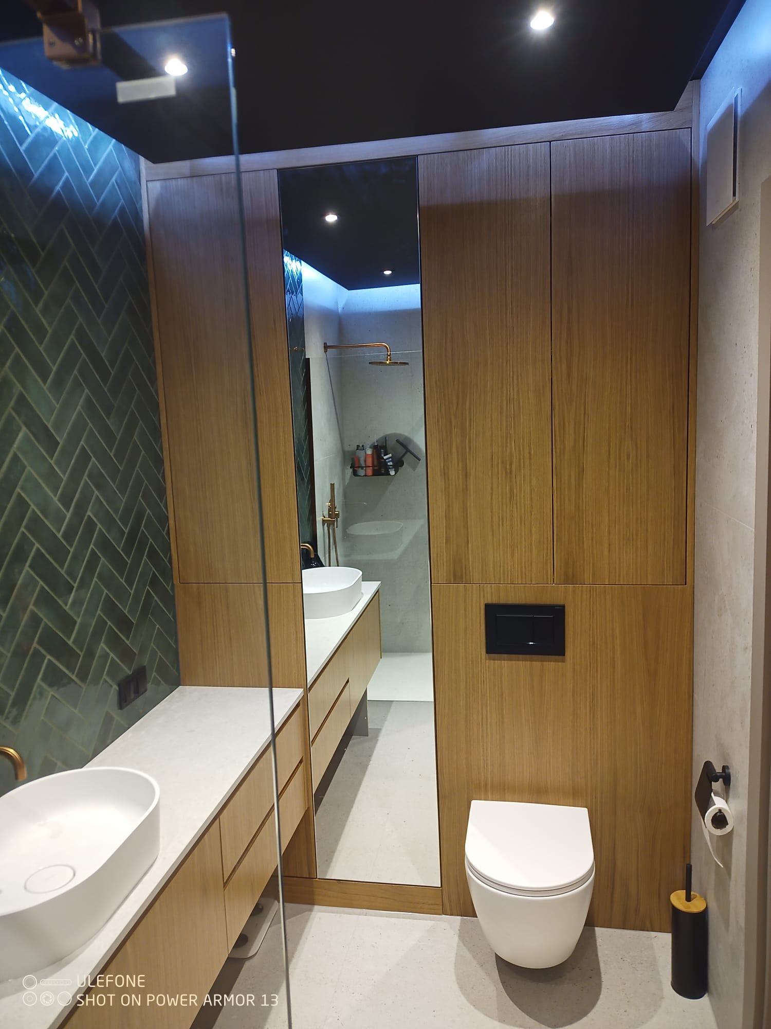 Nowoczesna łazienka z zieloną, ułożoną w jodełkę glazurą, drewnianymi szafkami i białym blatem z umywalką. W lustrze widoczny prysznic z deszczownicą.