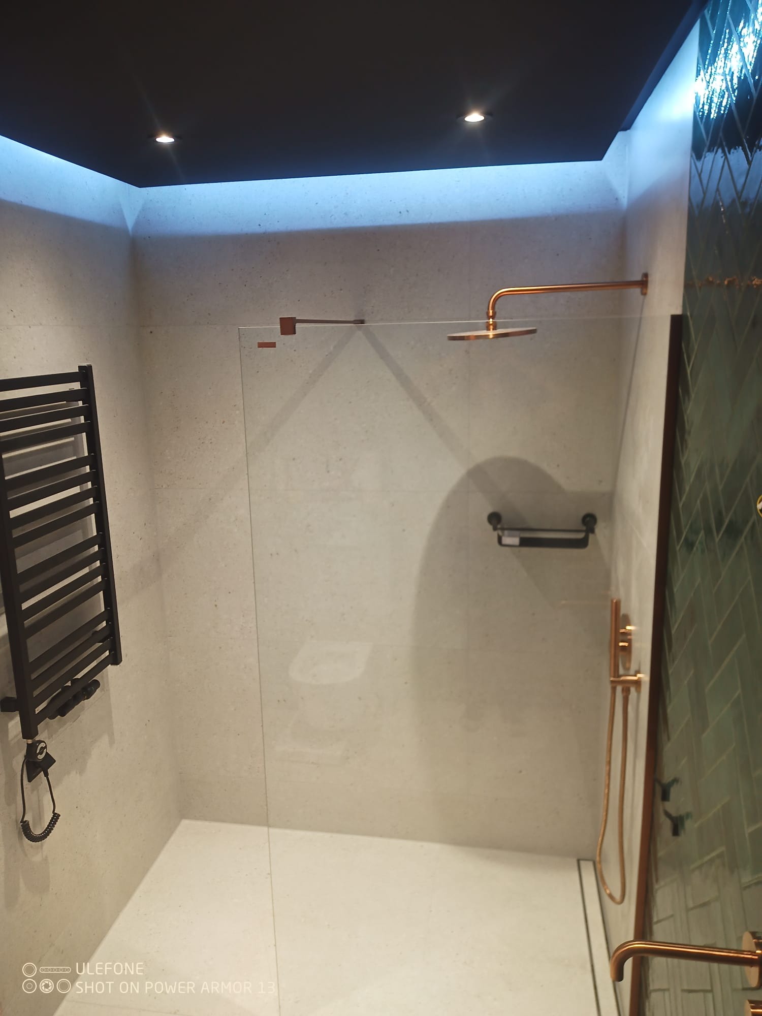 Nowoczesna kabina prysznicowa walk-in z miedzianą armaturą, grzejnikiem drabinkowym i oświetleniem LED w wykończonej łazience. Widoczne płytki na ścianach i podłodze.