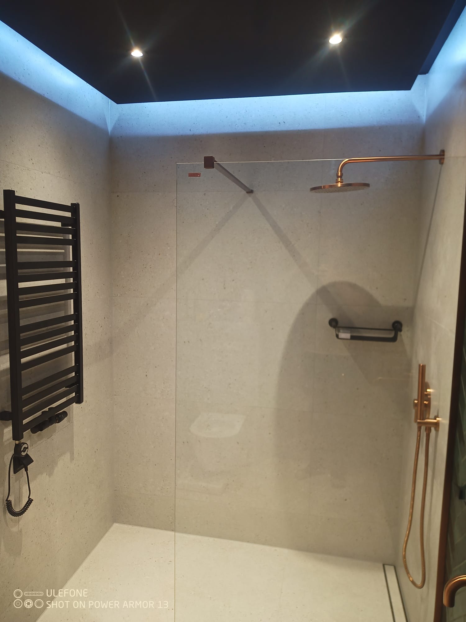 Nowoczesna kabina prysznicowa walk-in z deszczownicą w odcieniach miedzi, grzejnikiem drabinkowym i oświetleniem LED. Minimalistyczny design z jasnymi płytkami.