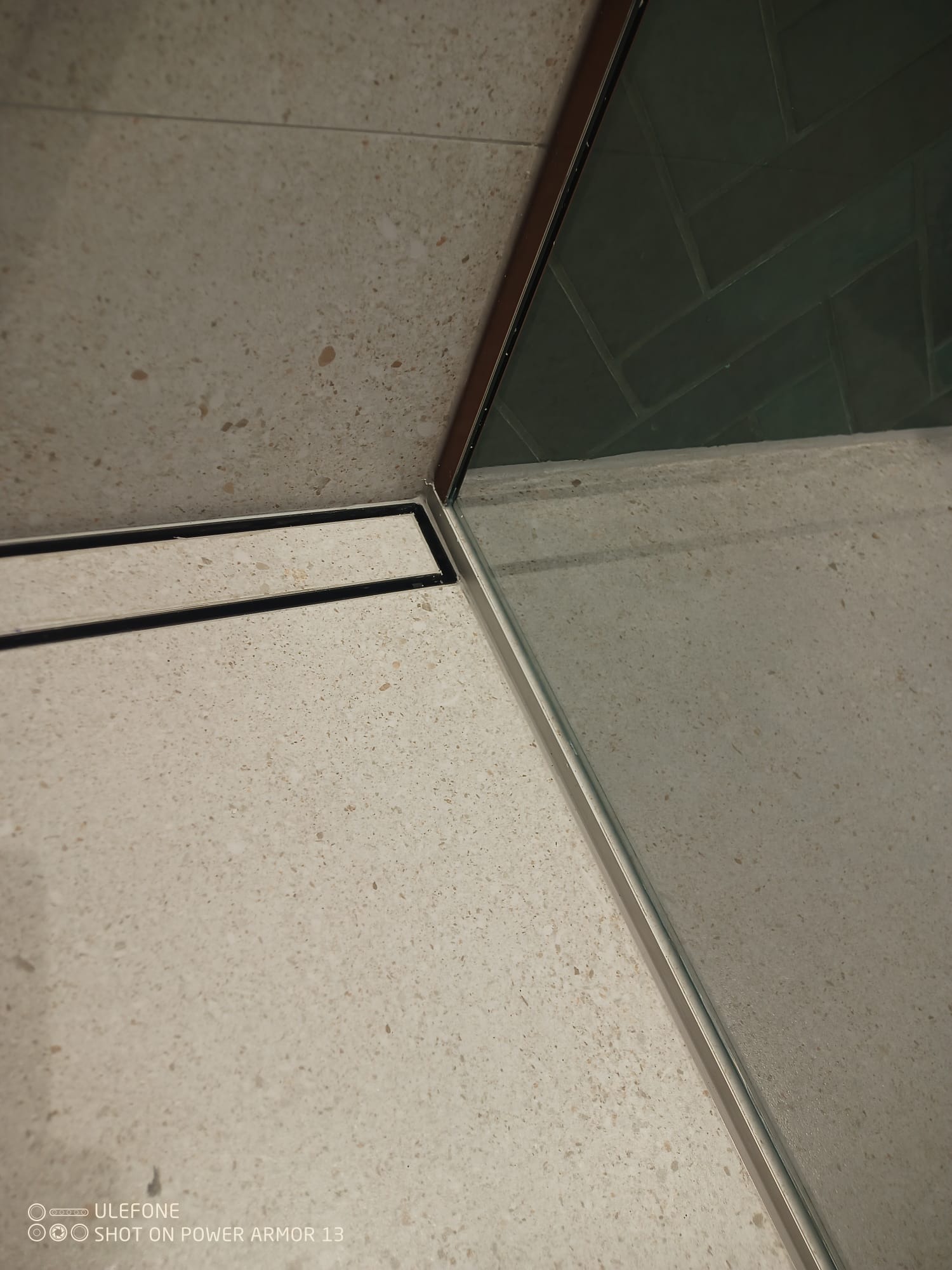 Beżowe płytki podłogowe z odpływem liniowym i szklaną kabiną prysznicową z ciemnozielonymi płytkami ułożonymi w jodełkę.