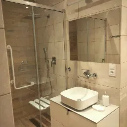 Jasna łazienka z kabiną prysznicową wyposażoną w siedzisko i uchwyt dla osób z ograniczeniami ruchowymi, umywalką nablatową na szafce z szufladami i lustrem z oświetleniem.