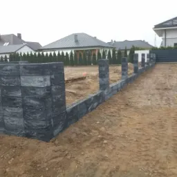 Wznoszenie muru oporowego z szarych, betonowych bloczków w surowym terenie budowy, widoczne fundamenty i przygotowany wykop pod dalszą konstrukcję, w tle domy jednorodzinne i rząd tuj.