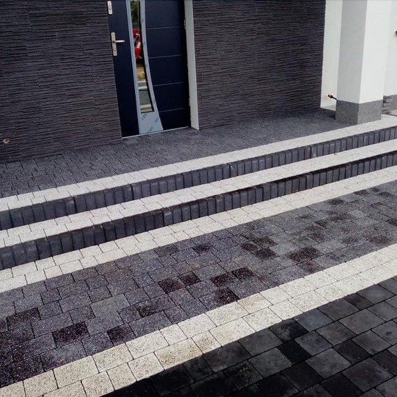 Schody wejściowe do domu wyłożone kostką w odcieniach szarości i bieli, z ciemną elewacją i drzwiami w tle.