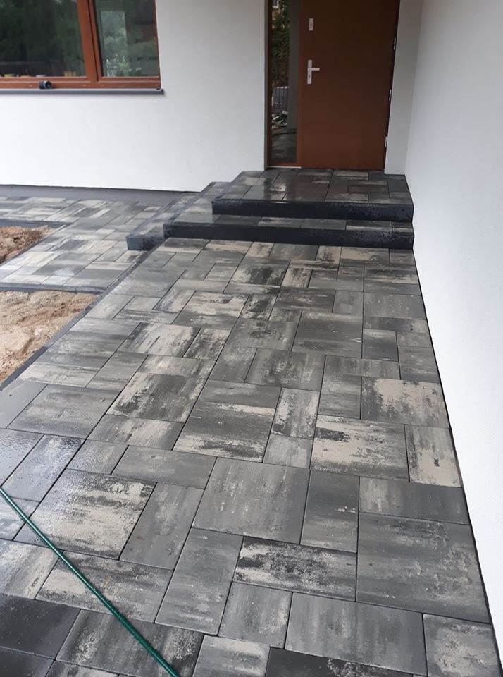 Ułożona szara kostka brukowa prowadząca do wejścia domu z brązowymi drzwiami, widoczne trzy stopnie z kostki, fragment zielonego węża ogrodowego w lewym dolnym rogu.